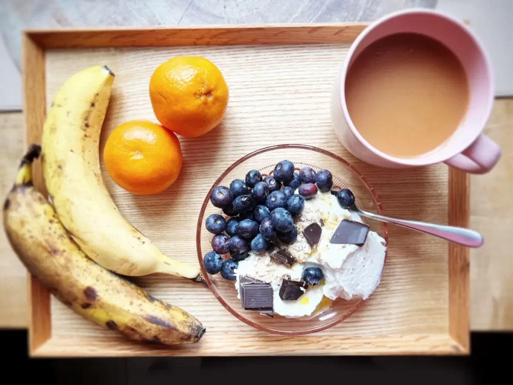 Fit śniadanie na tacy z miseczki jogurtu z borówkami, bananów i dwóch mandarynek i kawy. Obrazek w artykule 4 pomysły na szybkie fit śniadanie, które przygotujesz w mgnieniu oka