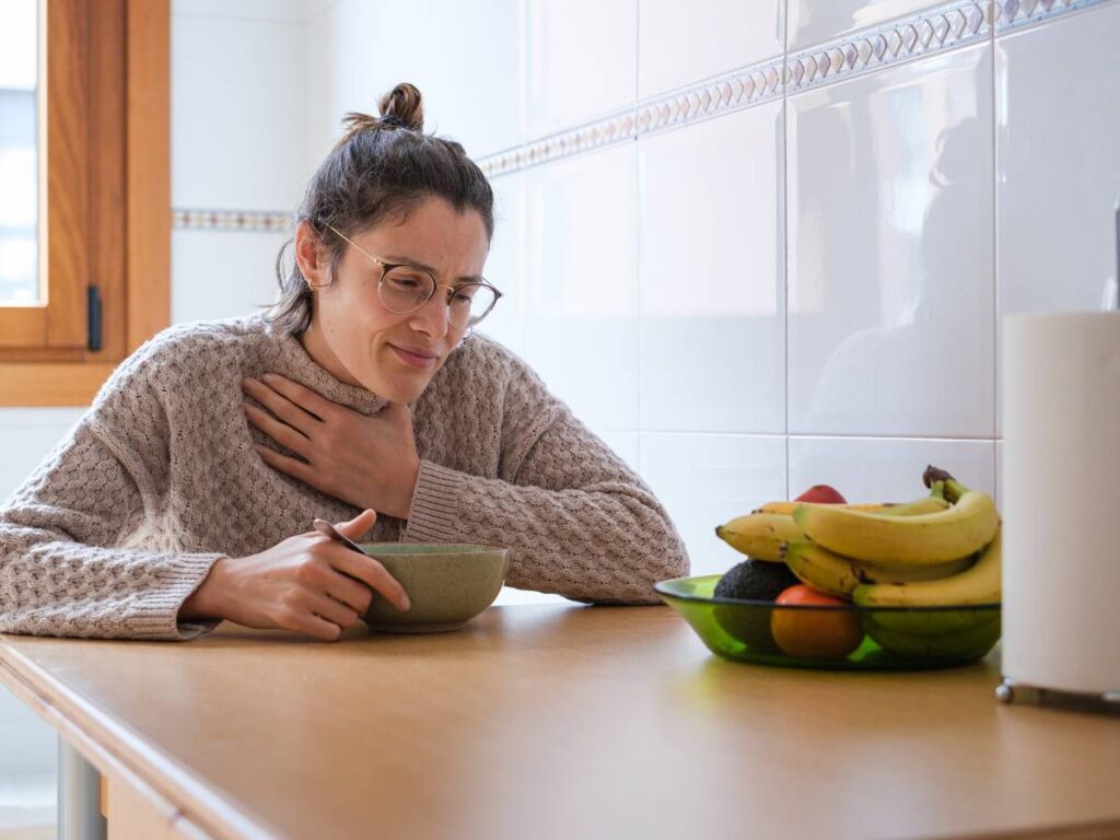 Kobieta przy stole jej coś z miseczki i owoce obok. Obrazek w artykule Doradzamy, kiedy jeść śniadanie, jeśli chcesz wdrożyć zdrowe nawyki żywieniowe