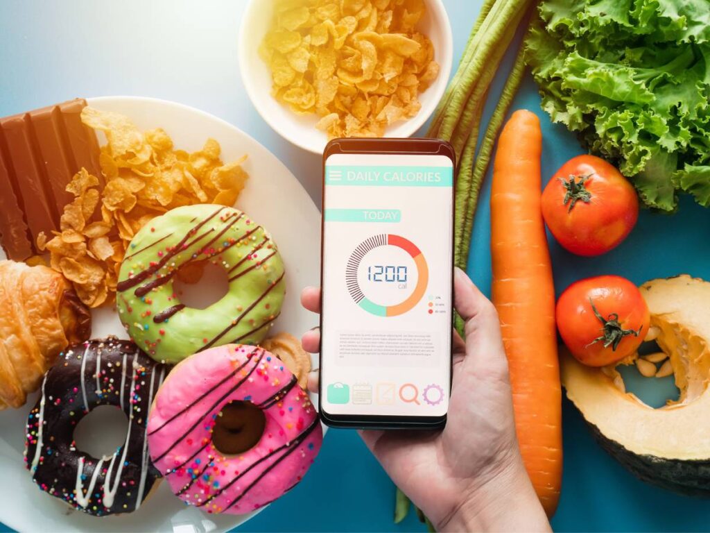 Produkty na stole: warzywa, marchewka, donuty i owsianka i pośrodku smartfon z otwartą aplikacją. Obrazek w artykule TOP 4 przepisy na wysokokaloryczne śniadanie, dla tych, którzy dużo spalają w ciągu dnia