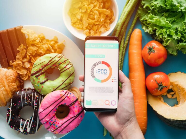 Produkty na stole: warzywa, marchewka, donuty i owsianka i pośrodku smartfon z otwartą aplikacją. Obrazek w artykule TOP 4 przepisy na wysokokaloryczne śniadanie, dla tych, którzy dużo spalają w ciągu dnia
