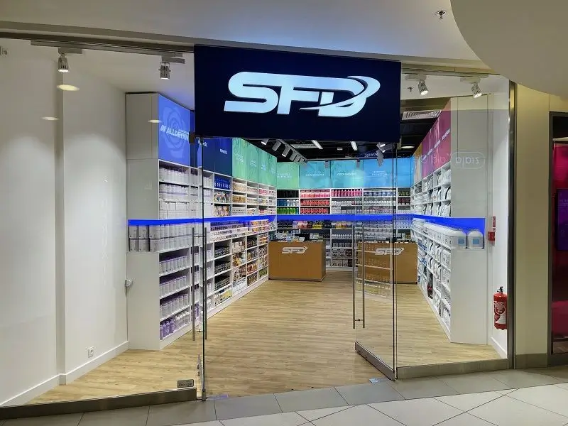 Zdjęcie przedstawia sklep marki SFD, który zajmuje się sprzedażą suplementów diety, odżywek oraz innych produktów związanych ze zdrowym stylem życia i sportem. Wejście do sklepu jest przeszklone, z dużym logo „SFD” umieszczonym na czarnym tle nad drzwiami. Wnętrze sklepu jest jasne i dobrze oświetlone, z kolorowymi regałami pełnymi produktów. Dominują jasne barwy, a podłoga wyłożona jest drewnopodobnym materiałem. W centralnej części sklepu widoczna jest lada z logo SFD. Po prawej stronie znajduje się gaśnica umieszczona na ścianie.