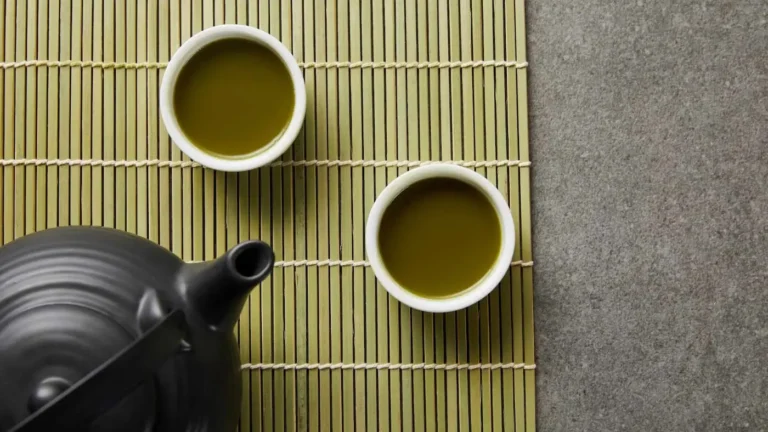 Zdjęcie przedstawia ceremonię picia herbaty w stylu azjatyckim, prawdopodobnie japońskim. W kadrze widoczny jest czarny ceramiczny czajnik oraz dwie małe filiżanki wypełnione zieloną herbatą, ustawione na bambusowej macie. Szczegóły obrazu: • Czajnik: • Matowy, czarny, o tradycyjnym, minimalistycznym kształcie. • Widoczna zakrzywiona wylewka i uchwyt, charakterystyczne dla japońskich imbryków Kyūsu, często używanych do parzenia herbaty sencha lub matcha. • Filiżanki: • Dwie małe, białe, bez uszu, co jest typowe dla tradycyjnej japońskiej i chińskiej kultury picia herbaty. • Wypełnione jasnozielonym naparem, prawdopodobnie zieloną herbatą o głębokim aromacie. • Mata bambusowa: • Jasnozielona, spleciona sznurkiem, służy jako podkładka i dodaje naturalnego, tradycyjnego charakteru. • Tło: • Szara, kamienna powierzchnia, która kontrastuje z ciepłymi tonami bambusa i herbaty. Atmosfera i przekaz: Zdjęcie oddaje spokój, harmonię i rytuał parzenia herbaty, co jest istotnym elementem kultury Japonii i Chin. Minimalistyczna kompozycja i staranny układ przedmiotów podkreślają zen i prostotę.