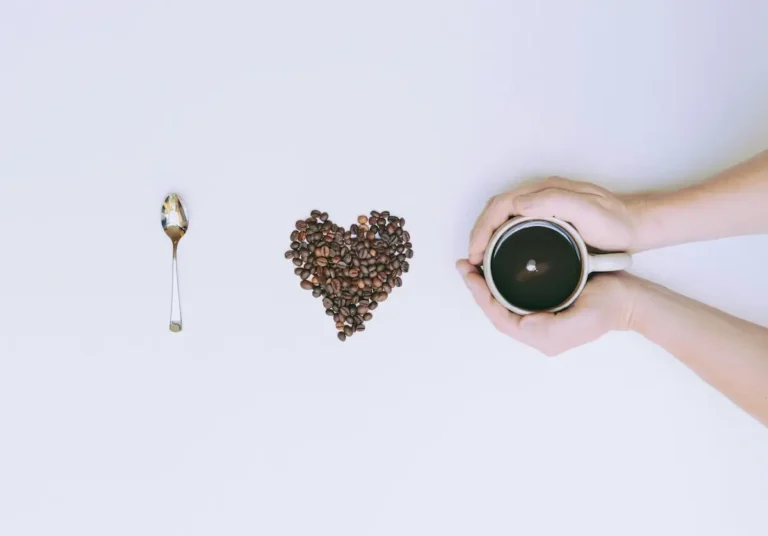 Symboliczna kompozycja przedstawiająca miłość do kawy – po lewej łyżeczka, pośrodku serce ułożone z ziaren kawy, po prawej filiżanka czarnej kawy trzymana w dwóch dłoniach na jasnym tle.