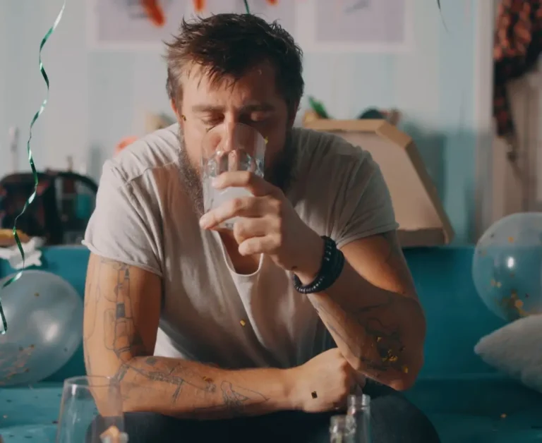 Zmęczony mężczyzna z tatuażami siedzi na kanapie w otoczeniu balonów i resztek po imprezie, z zamkniętymi oczami pije wodę z przezroczystej szklanki – wygląda na wyczerpanego i odwodnionego, co symbolizuje dolegliwości związane z kacem.